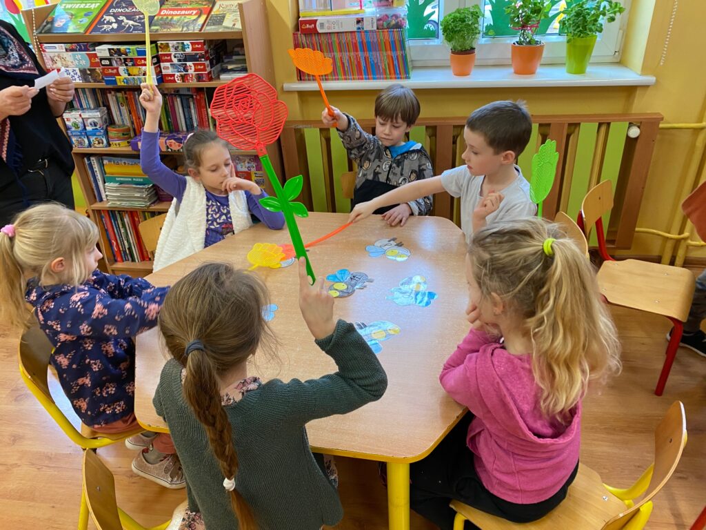 Przy 2 stolikach siedzą dzieci, trzymające klapki na muchy w rękach. Na stole przyklejone są muchy z zapisanymi działaniami.