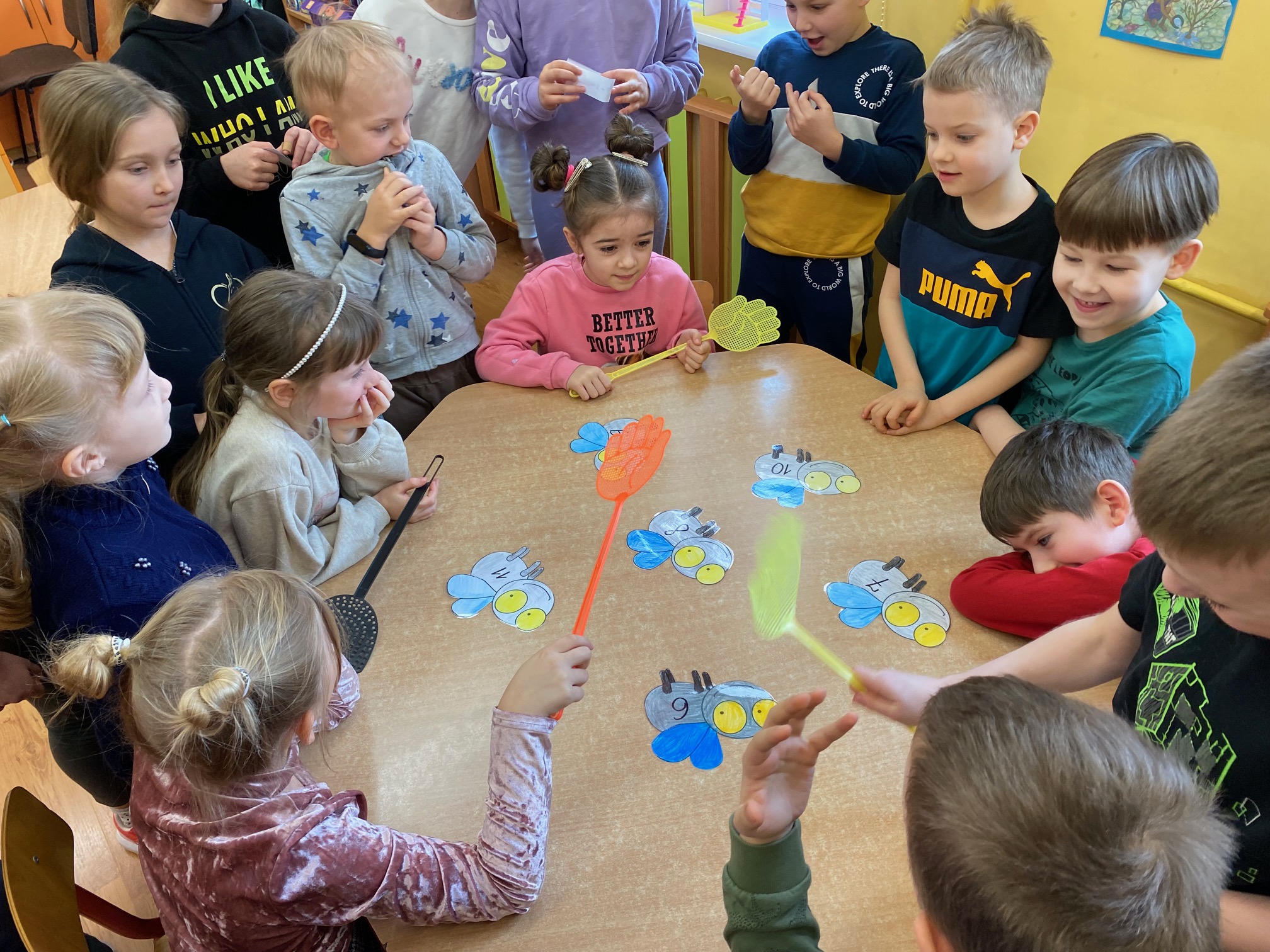 Przy stoliku siedzą dzieci, trzymające klapki na muchy w rękach. Na stole przyklejone są muchy z zapisanymi działaniami. Przy stole stoją również dzieci z karteczkami z działaniami.