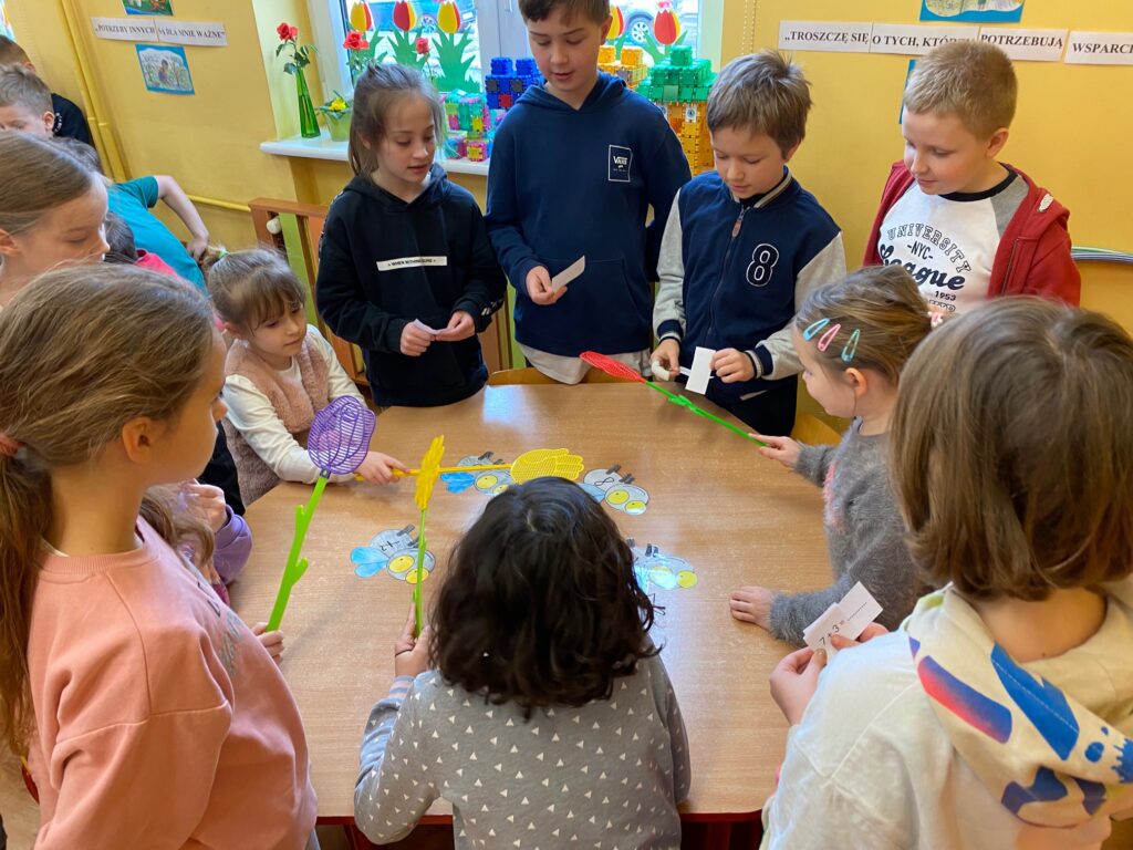 Przy stoliku siedzą dzieci, trzymające klapki na muchy w rękach. Na stole przyklejone są muchy z zapisanymi działaniami. Przy stole stoją również dzieci z karteczkami z działaniami.