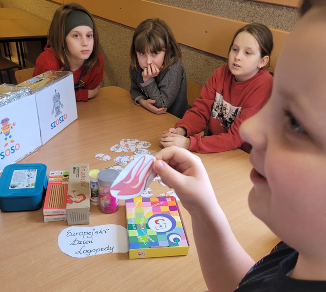 Na pierwszym planie widać twarz chłopca, który w trzyma w dłoni małą karteczkę z obrazkiem butów na szpilce. Chłopiec jest zaciekawiony. W tle widać troje dzieci siedzących przy stole. Na stole leżą pomoce dydaktyczne do zajęć- pudełka z grami, napis "Europejski Dzień Logopedy", dwa pudełka z obrazkami robotów "SZOSZO' i "SOSO".