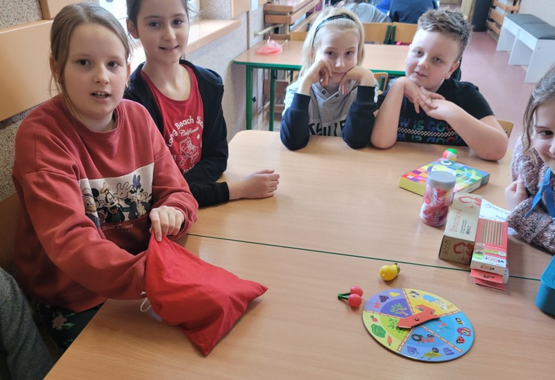 Na zdjęciu widać grupę dzieci siedzących przy stole. Na stole leżą pomoce dydaktyczne do zajęć, pudełka, kolorowe koło fortuny oraz czerwony woreczek, z którego dziewczynka siedząca po lewej stronie próbuje coś wyciągnąć.