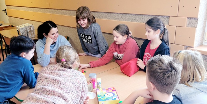 Na zdjęciu widać grupę dzieci siedzących przy wspólnym stole. Między dziećmi siedzi ciemnowłosa kobieta. Jedna z dziewczynek stoi i pochyla się nad stołem. Na stole widać pomoce dydaktyczne do zajęć.