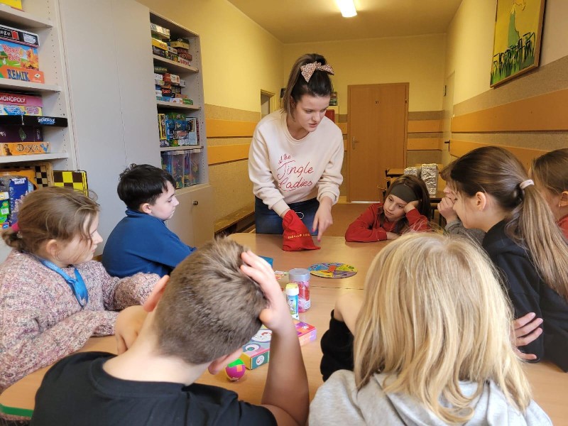 Na zdjęciu znajdują się uczniowie, którzy siedzą przy stole. Na przeciwko uczniów stoi kobieta w kremowej bluzie, upiętym kucyku na głowie i kokardą we włosach. Wzrok uczniów skupiony jest na czerwonym worku, w którym kobieta trzyma rękę.