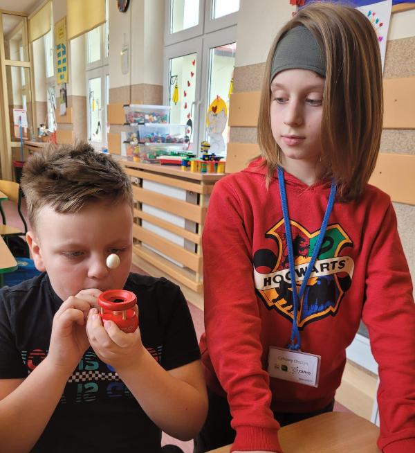Na zdjęciu znajduje się dwóch chłopców. Chłopiec z lewej strony siedzi na krześle, ma na sobie czarną koszulkę. Dmucha w dmuchajkę za pomocą słomki. Biała kulka unosi się w powietrzu. Z prawej strony stoi chłopiec ubrany w czerwoną bluzę. Jego wzrok skierowany jest na chłopca, który znajduje się z jego prawej strony.