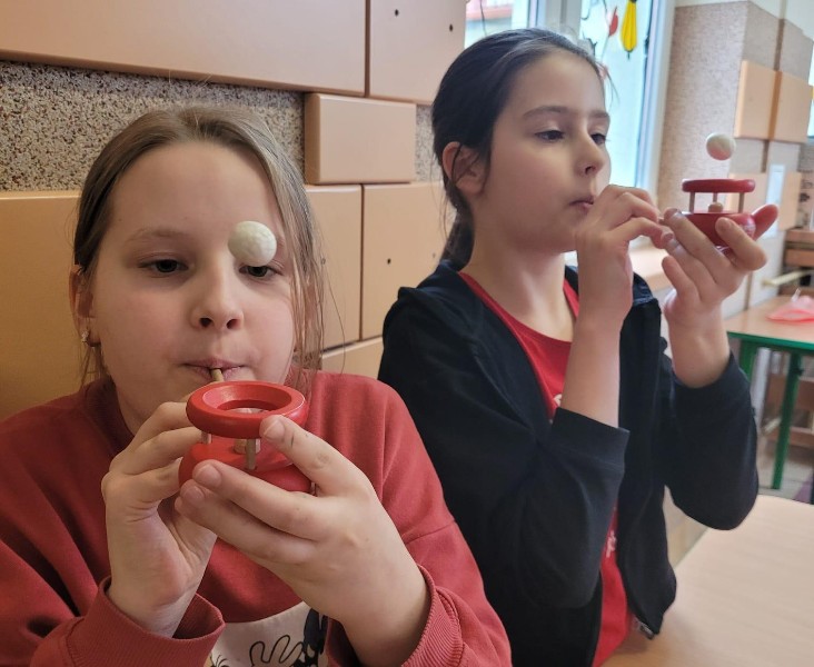 Na zdjęciu widać dwie dziewczynki, które siedzą obok siebie przy stole. Dziewczynka znajdująca się po lewej stronie ma blond włosy i ubrana jest w czerwoną bluzę. Dziewczynka obok ma brązowy kolor włosów i ubrana jest w czerwoną bluzkę i czarny sweterek. Obie dziewczynki dmuchają za pomocą słomki dmuchajki. Białe kulki unoszą się w powietrzu.