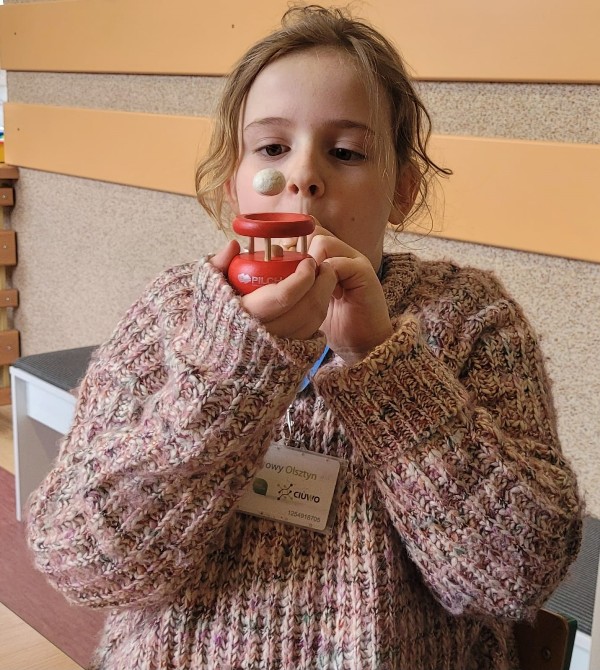 Na zdjęciu znajduje się dziewczynka o blond włosach. Ubrana jest w sweter w jasnych kolorach. W dłoniach trzyma dmuchajkę, w którą dmucha za pomocą słomki. Biała kulka unosi się w powietrzu.