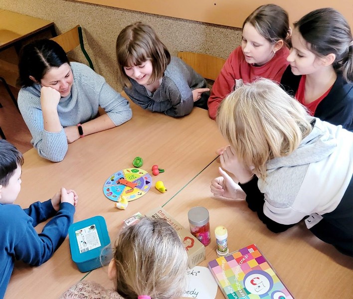 Na zdjęciu znajdują się uczniowie, którzy siedzą w kręgu. Wśród uczniów znajduje się dorosła, ciemnowłosa kobieta, która podpiera głowę ręką. Wszyscy mają zwrócony wzrok na ławkę, na której znajduje się kolorowe koło fortuny oraz gumowe owoce: wiśnia, sałata, gruszka.