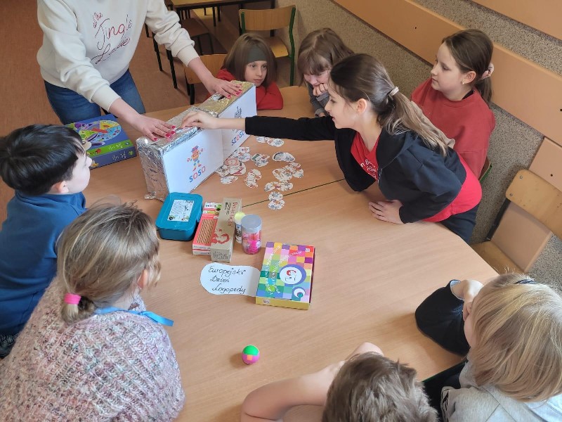 Na zdjęciu znajdują się uczniowie, którzy siedzą w kręgu przy stole. Na stole leżą pomoce logopedyczne: piórka, dmuchajki, słomki, pudełko. Ciemnowłosa dziewczynka, ubrana w czerwoną bluzkę i czarny sweter wkłada prawą ręką wylosowany obrazek, do kartonika z napisem „SOSO”.