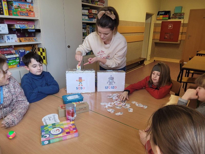 Na pierwszym planie znajduje się kobieta ubrana w kremową bluzę Na głowie ma upięty kucyk i kokardę. W prawej dłoni trzyma obrazek, który przedstawia ananasa. Palcem wskazującym lewej ręki pokazuje na obrazek, który wkłada do kartonu z napisem „SOSO”. Obok kartonów znajdują się pomoce logopedyczne. Chłopiec w czerwonej bluzie losuje obrazek.