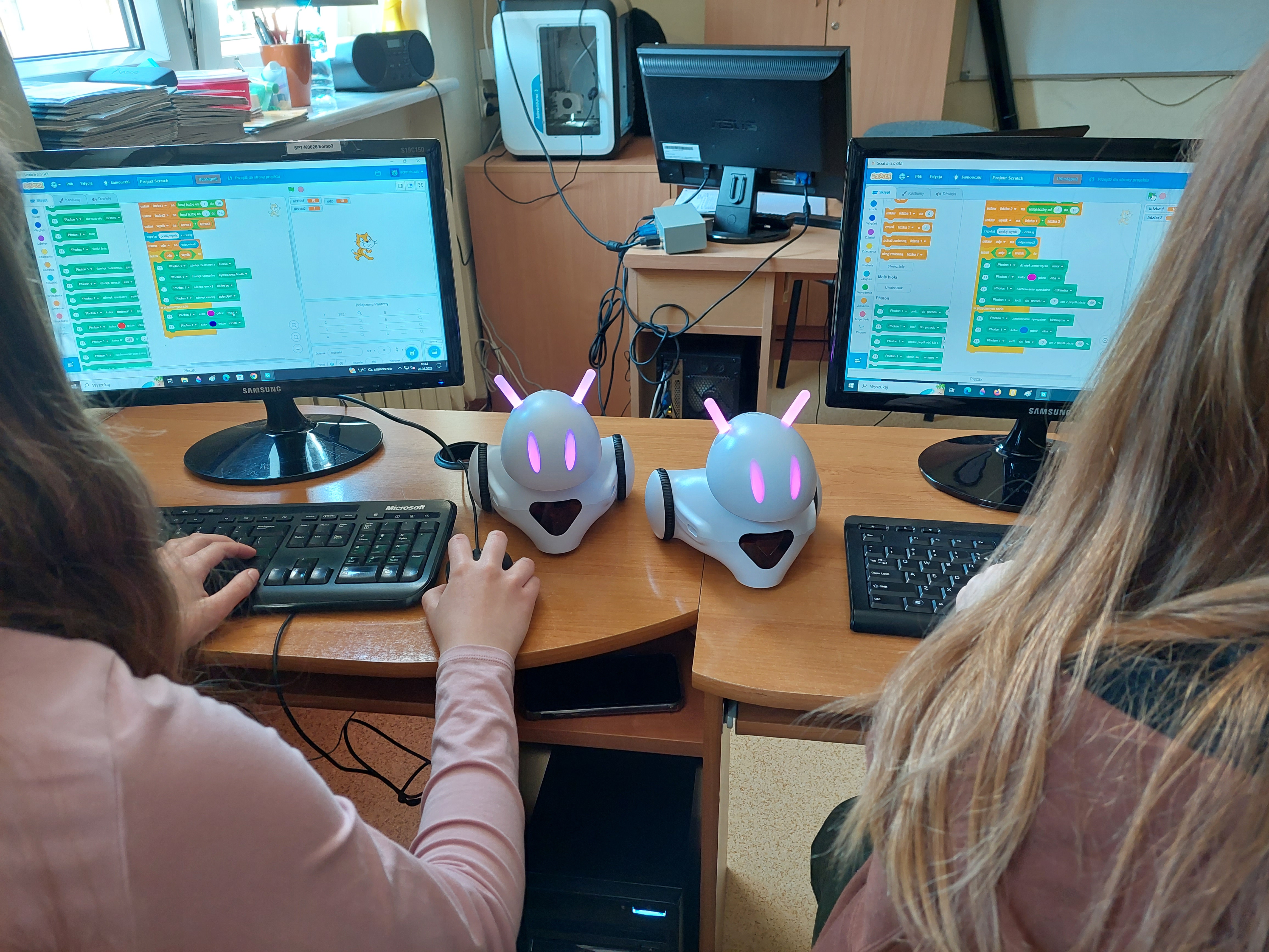 Dwa białe roboty. Każdy z nich ma kolorowe diody. Po bokach widoczne monitory z fragmentem kodu oraz pracujące nad nim uczennice.