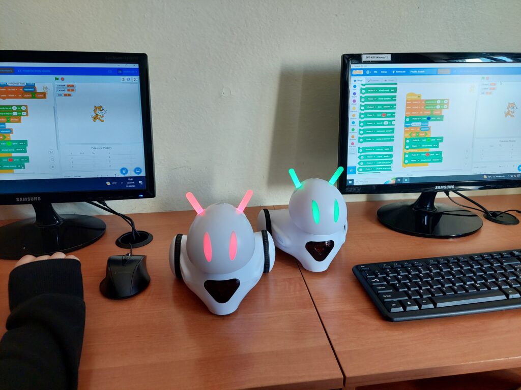 Dwa białe roboty. Każdy z nich ma kolorowe diody. Po bokach widoczne monitory z fragmentem kodu.