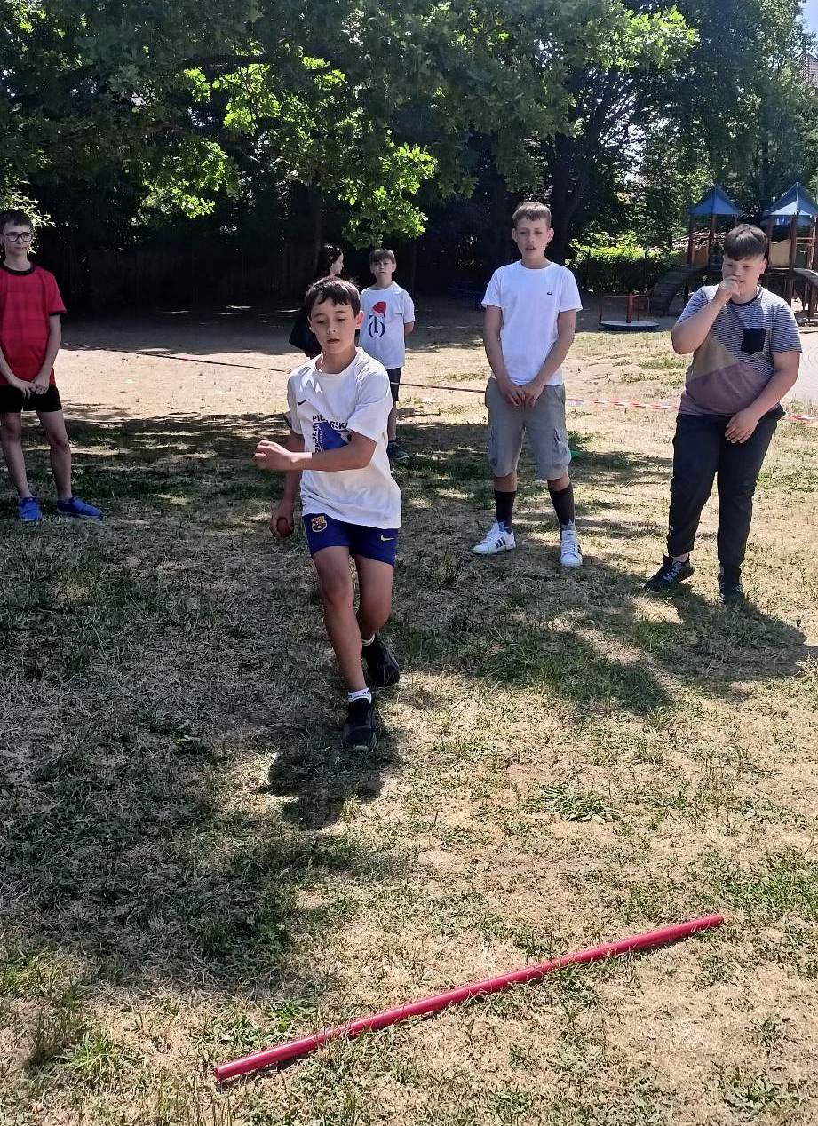 Zdjęcie przedstawia chłopca wykonującego rzut piłeczką .Ubranego w struj sportowy Za uczniem stoi trzech chłopców przyglądających się zawodnikowi.