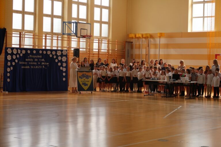 Na zdjęciu znajduje się kobieta z mikrofonem białej sukience stojąca na tle niebieskiej ścianki ozdobionej napisem i białymi kwiatami. Przed kobietą stoi tablica ze znakiem szkoły. Kobieta zwrócona jest twarzą w stronę grupy dzieci ubranych na biało czarno za dziećmi stoją dorośli.