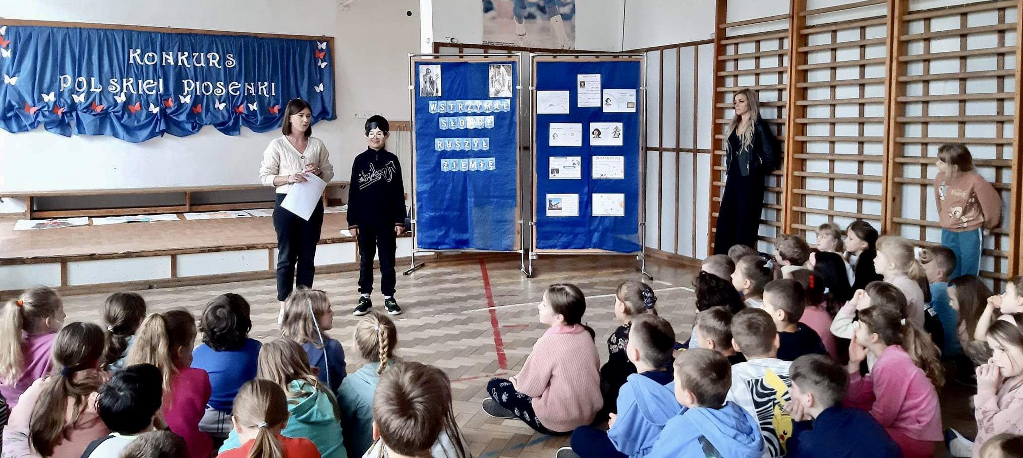Dzieci siedzą na podłodze. Nauczyciel prowadzi spotkanie. Chłopiec w masce Mikołaja Kopernika stoi na środku sali.