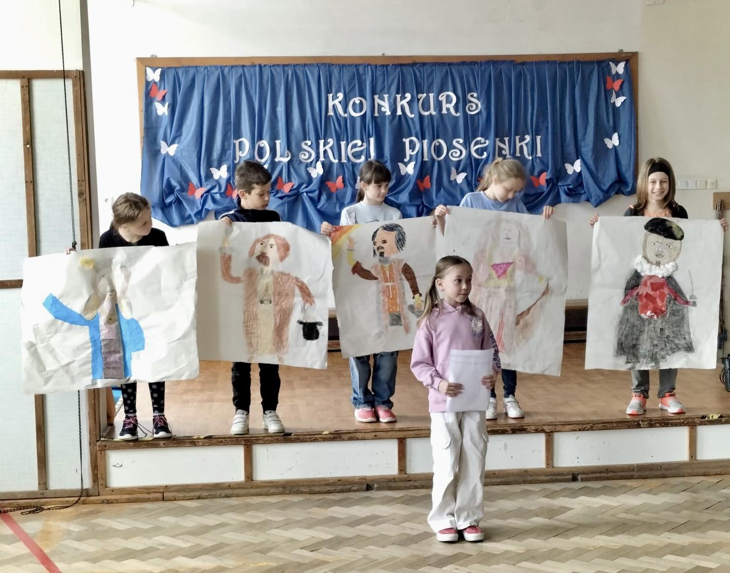 Dzieci trzymają w rękach duże kartki na których narysowane są średniowieczne stroje. Dziewczynka trzyma w ręku kartkę.