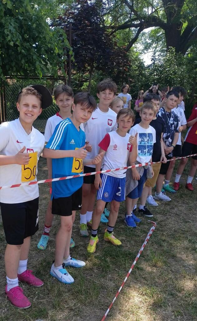 Zdjęcie przedstawia grupę chłopców ubranych w stroje sportowe, krótkie spodenki i koszulki na krótki rękaw stojących za biało czerwona taśmą.