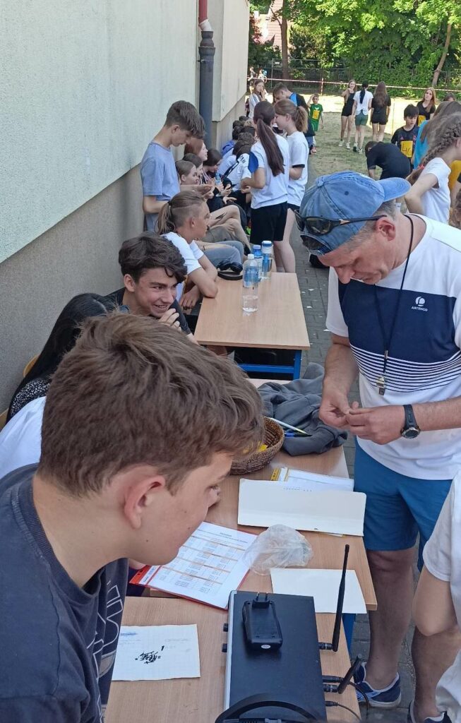 Na zdjęciu znajdują się stoliki ustawione pod ścianą na podwórku. Wokół stolików stoją lub siedzą uczniowie. Na pierwszym planie znajduje się mężczyzna ubrany na sportowo z czapka na Glowie daszkiem odwróconym do tyłu na czapce ma okulary słoneczne. Druga postać pierwszego planu to chłopiec którego widać tylko głowę część twarzy i ucho.