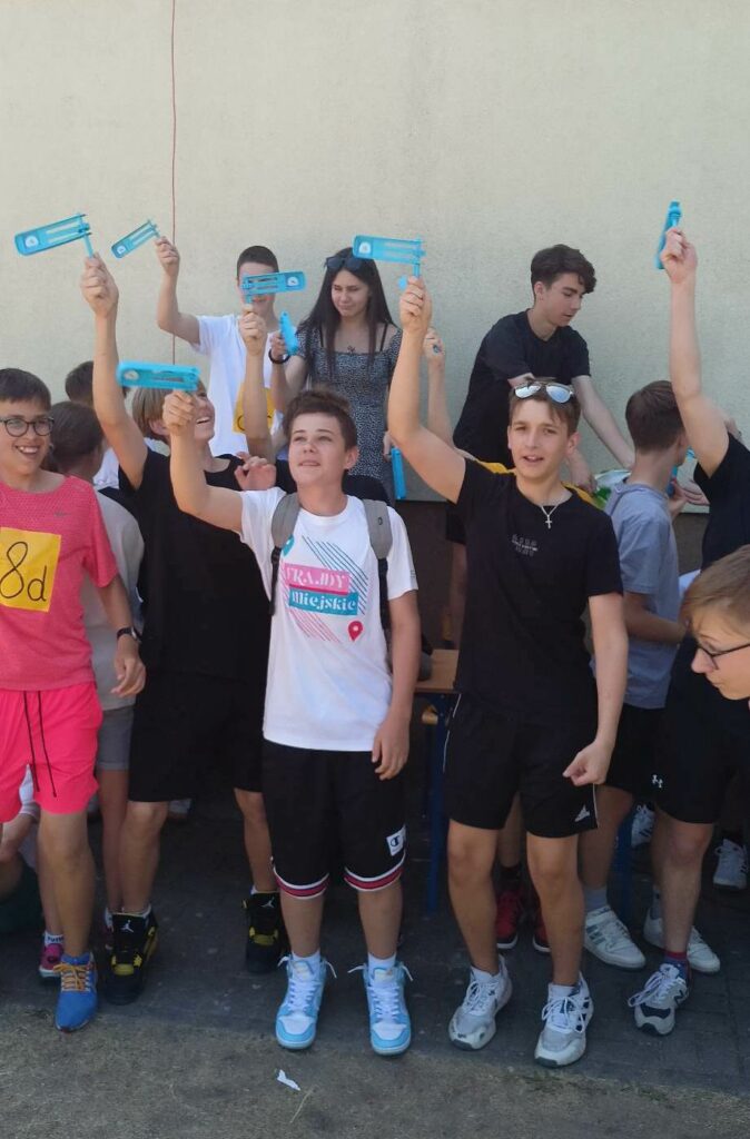 Zdjęcie przedstawia grupę uczniów chłopców i dziewczynę trzymających w rękach wyciągniętych w górę niebieskie grzechotki .