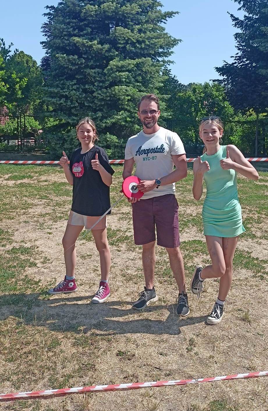 Zdjęcie przedstawia trzy osoby stojące na tle drzew i czerwono białej taśmy. Z Lewej strony dziewczyna w jasnych krótkich spodenkach i czarnej koszulce. Kolejna osoba to mężczyzna w stroju sportowym trzymający w rekach taśmę mierniczą. Trzecia osoba to dziewczyna w zielonej sukience. Dziewczęta trzymają kciuki obu rąk do góry.