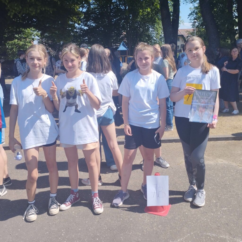 Grupa czterech dziewcząt na tle wielu osób. Dziewczęta ubrane w białe koszulki trzy z nich mają krótkie spodenki jedna długie. Dziewczyna w długich spodniach stoi z lewej strony zjecie i trzyma w dłoniach dyplom.