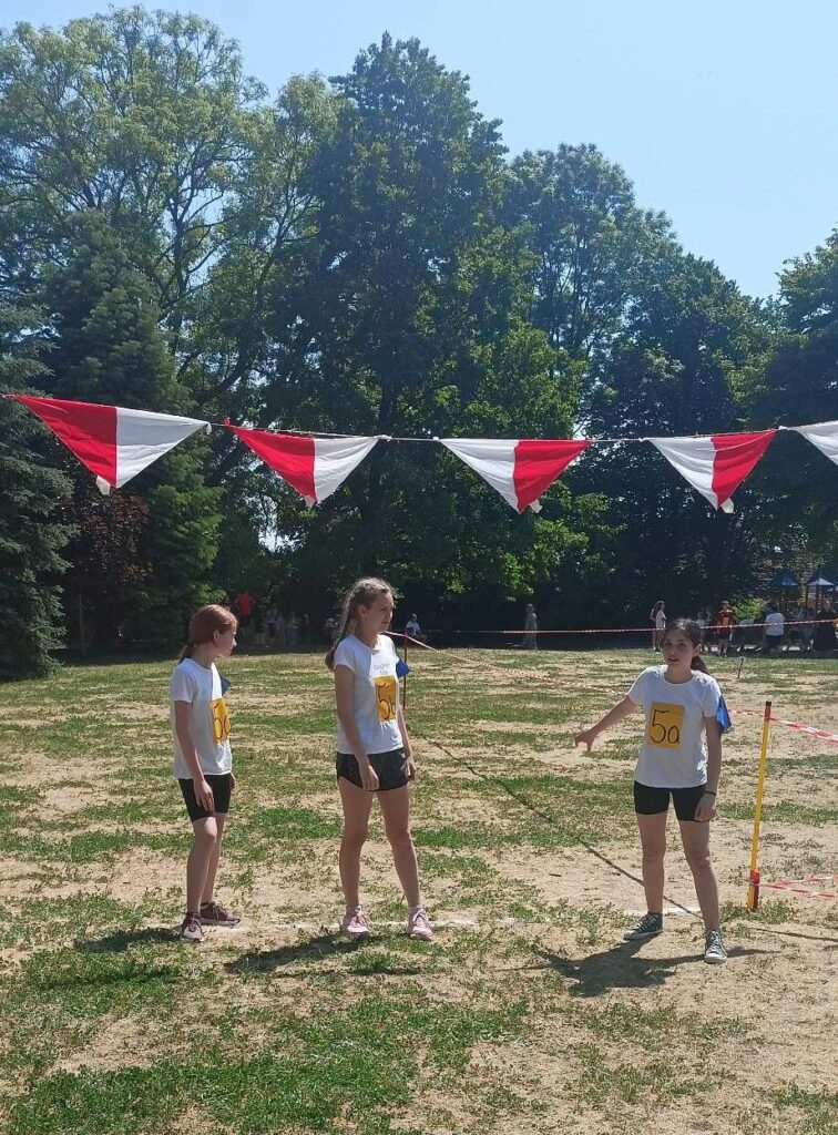 Zdjęcie przedstawia trzy dziewczyny ubrane w czarne krótkie spodenki i białe koszulki na których znajdują się przyklejone żółte naklejki z napisem 5a. Stoją na tle drzew nad nimi Wisza biało czerwone trójkątne girlandy.
