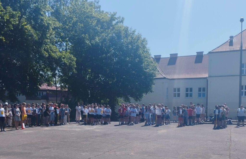 Zdjęcie przedstawia grupy uczniów stojących na boisku szkolnym ustawionych klasami.
