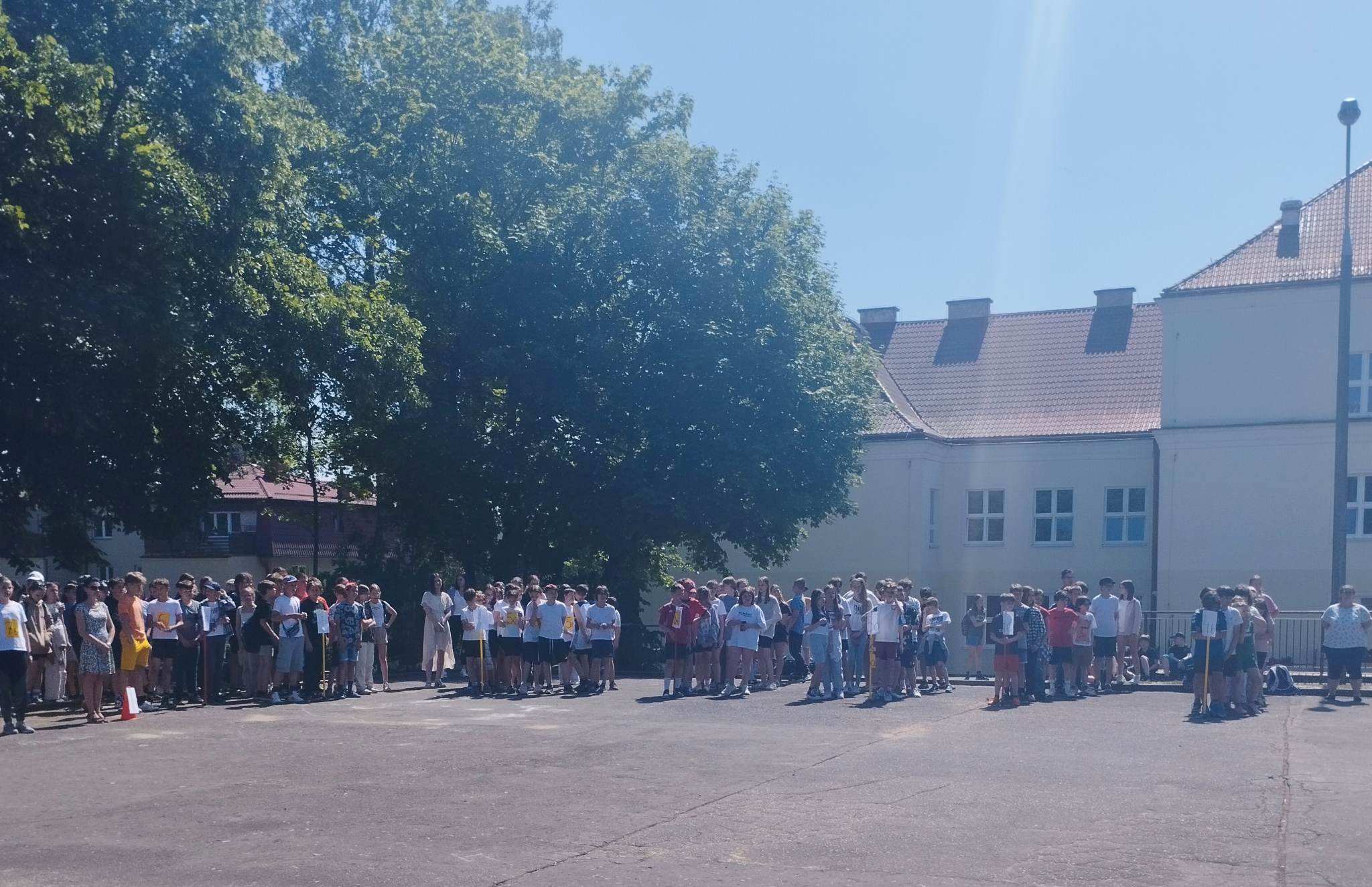 Zdjęcie przedstawia grupy uczniów stojących na boisku szkolnym ustawionych klasami.