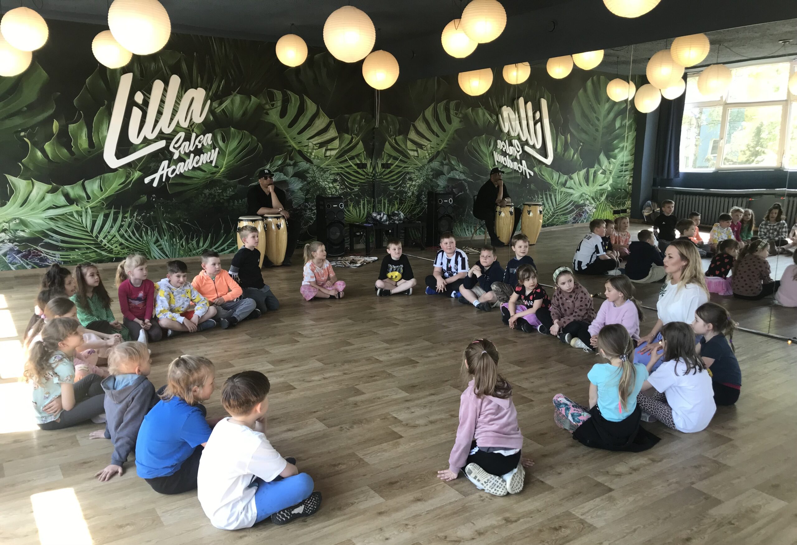 Dzieci siedzą w kole na drewnianej podłodze. Na ścianie widać napis Lilla Salsa Academy oraz tapetę, na której są zieone liście palmy. Przy kongach siedzi ciemnoskóry mężczyzna.
