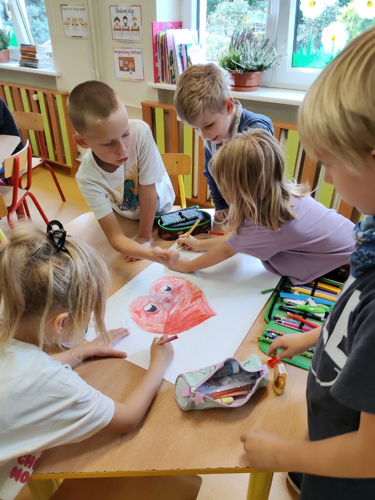na zdjęciu dzieci robią plakat z okazji dnia empatii. Dwóch chłopców i dwie dziewczynki. na stoliku duża kartka i piórnik.