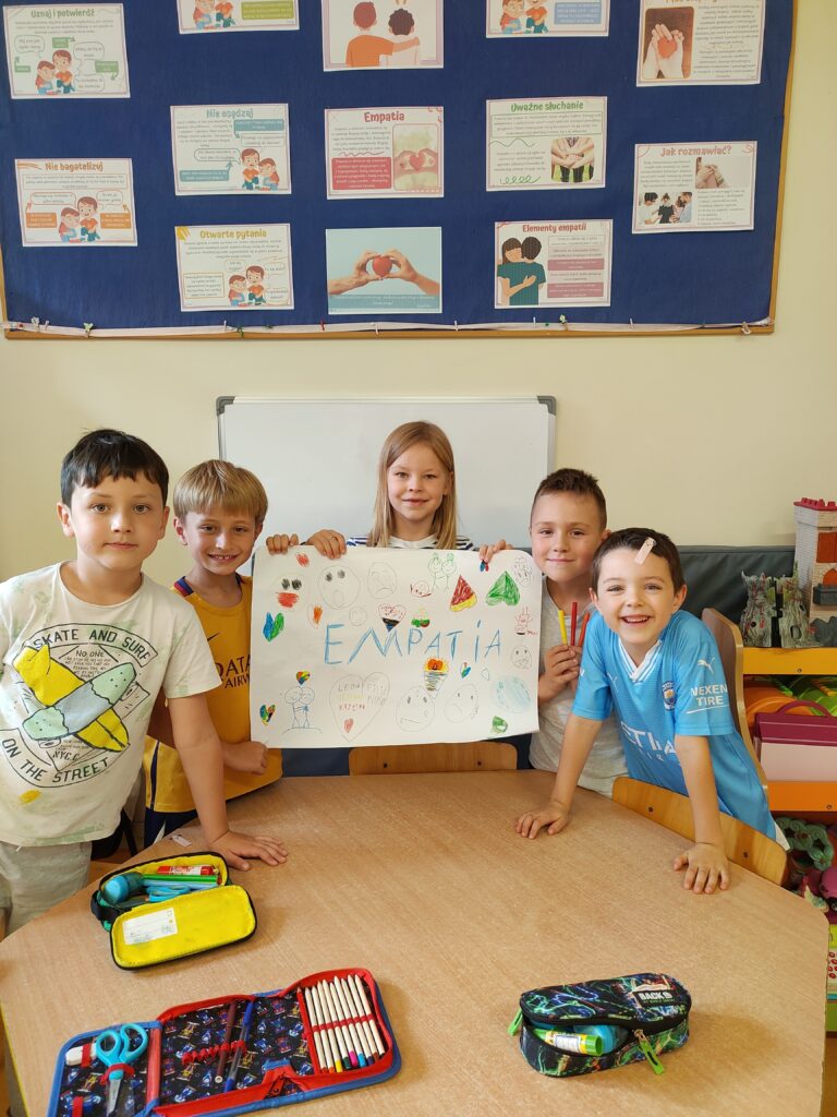 czterech chłopców i jedna dziewczynka stoją wokół stolika. dziewczynka trzyma w ręku plakat. Na środku plakatu niebieski napis empatia. w tle tablica scienna