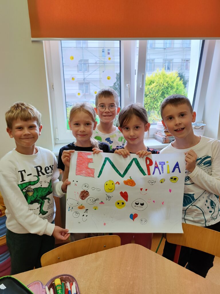 dwie dziewczynki i trzech chłopców stoją obok siebie i trzymają plakat na którym jest kolorowy napis empatia