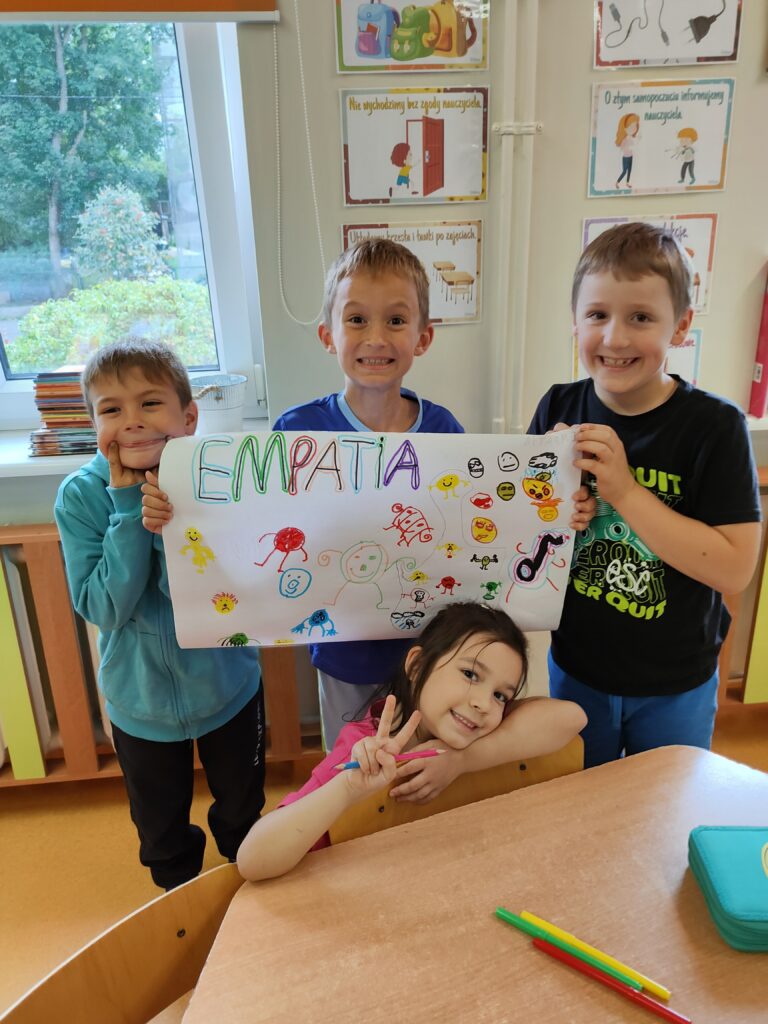 trzech chłopców stoi i prezentuje plakat który zrobili sami, na plakacie napis empatia i kolorowe stworki, w tle okno