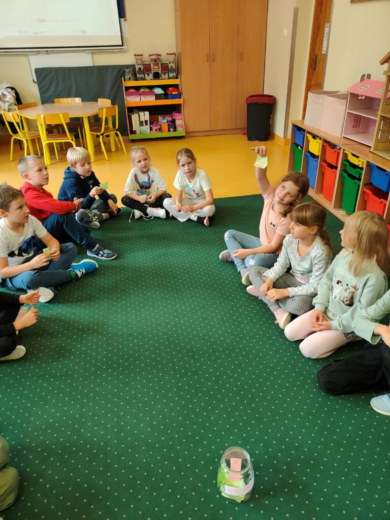 na zdjęciu wdać grupkę dzieci siedzących na zielonym dywanie. Dzieci siedzą w kręgu. Jedna z dziewczynek trzyma w ręku karteczkę.