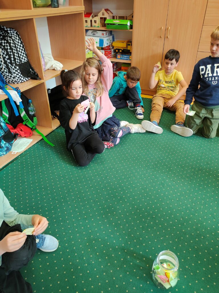 na zdjęciu widać dzieci siedzące na dywanie. Obok siebie dwie dziewczynki, jedna czyta karteczkę. Obok siedzą chłopcy. Jedna z dziewczynek podnosi rękę do góry