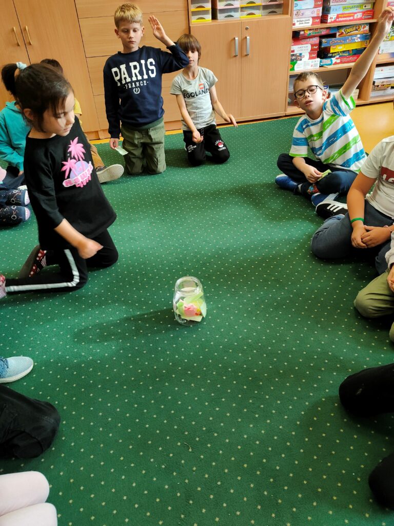 dzieci siedzą na dywanie w kręgu. Jedna dziewczynka wyciąga rękę by wylosować karteczkę ze słoika, który stoi na środku dywanu.