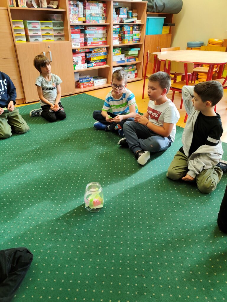dzieci siedzą na dywanie w kręgu. Po środku stoi duży słoik, w środku słoika karteczki. Siedzące dzieci, trzymają w ręku karteczki