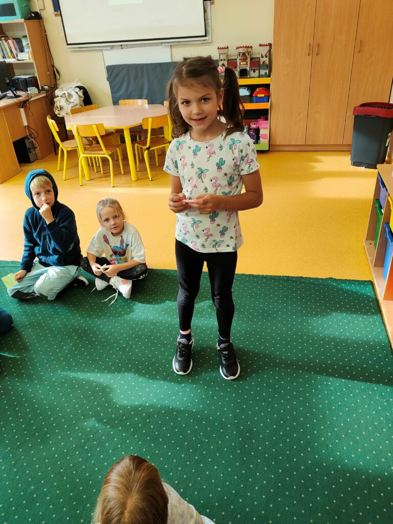 na zdjećiu stoi dziewczynka, w ciemnych włosach i kolorowej koszulce. trzyma w ręku karteczkę. za nią siedzą dzieci na dywanie. Dziewczynka i chłopiec. w Tle stolik.