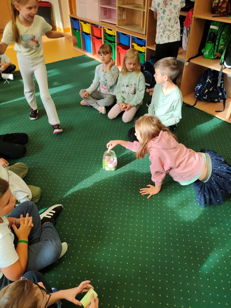 dzieci siedzą na dywanie w kręgu. Jedna dziewczynka wyciąga rękę by wylosować karteczkę ze słoika, który stoi na środku dywanu.. Podchodzi do słoika, też inna dziewczynka