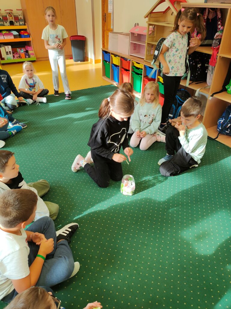 dzieci siedzą na dywanie w kręgu. Jedna dziewczynka wyciąga rękę by wylosować karteczkę ze słoika, który stoi na środku dywanu.