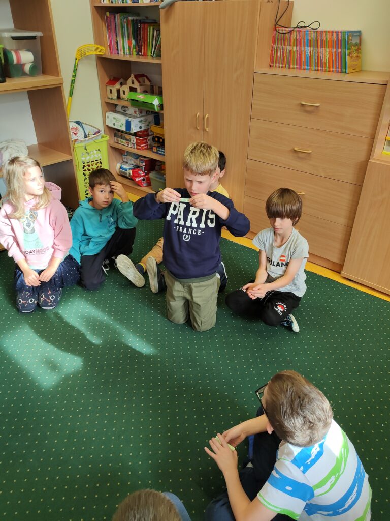 na dywanie siedzą dzieci. Jeden chłopiec klęczy. trzyma w ręku małą karteczkę