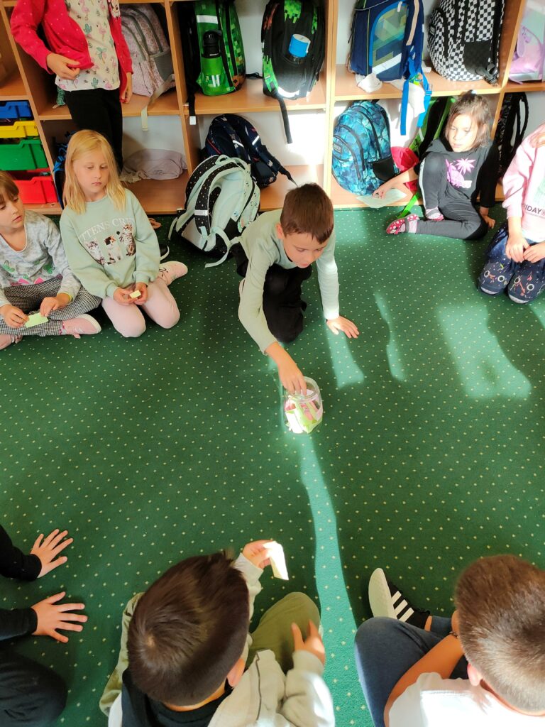 dzieci siedzą na dywanie w kręgu. Jeden chłopiec wyciąga rękę by wylosować karteczkę ze słoika, który stoi na środku dywanu.