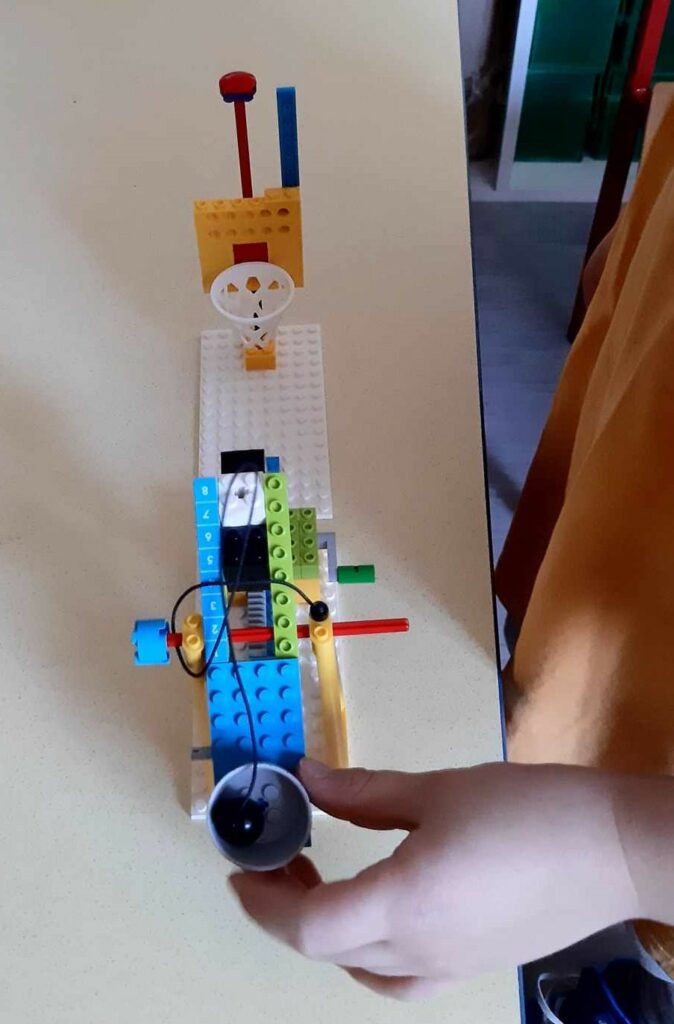 Zdjęcie przedstawia konstrukcję z klocków Lego ustawioną na jasnym stole. Ręka dziecka trzyma jedną część konstrukcji, która jest koszem z czarna kulką na sznurku, którą celuję w druga cześć konstrukcji, którą jest biały kosz na żółtej tablicy z klocków.