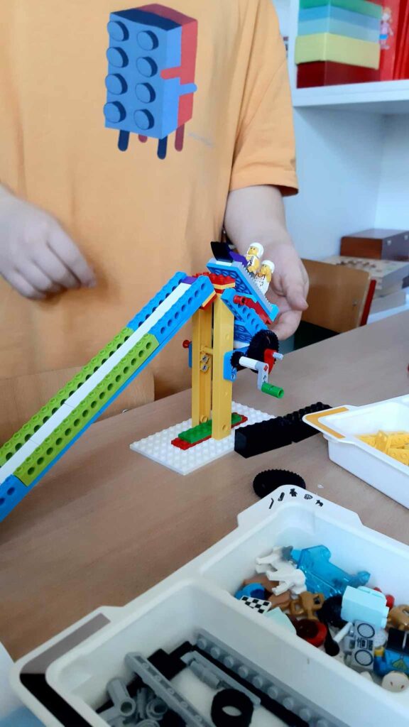 Zdjęcie przedstawia konstrukcję toru saneczkarskiego z klocków lego. Za konstrukcja jest postać dziecka trzymającego lego sanki z ludzikami umieszczając je na zjeżdżalni.