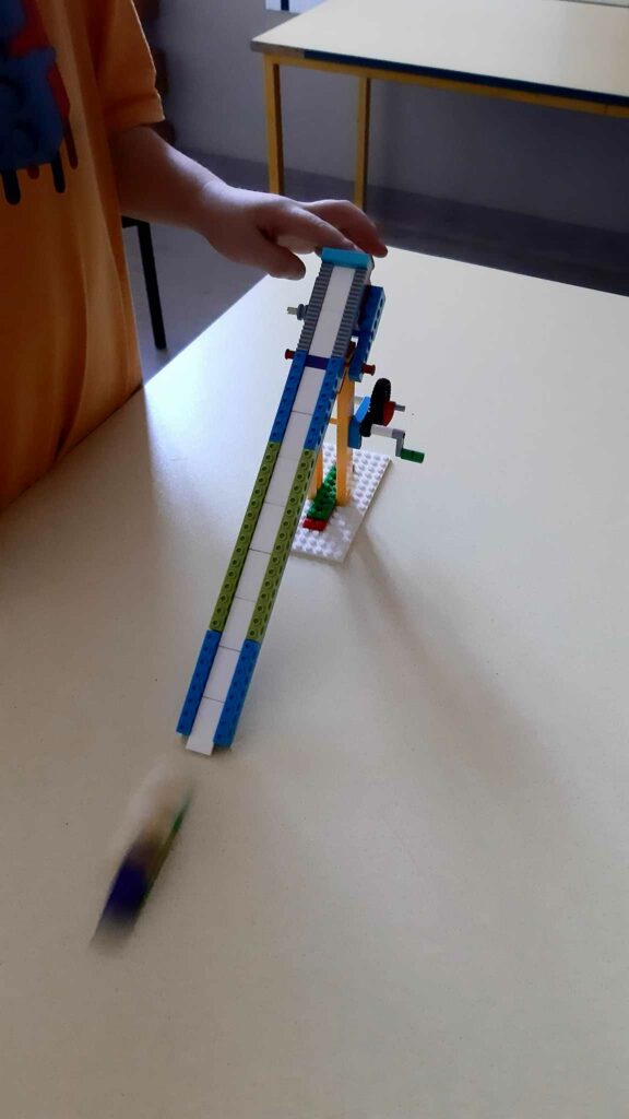 Na zdjęciu widać konstrukcję toru saneczkarskiego z Lego ustawionego na stole. Przy stole widać fragment ręki dziecka żółtą koszulkę.