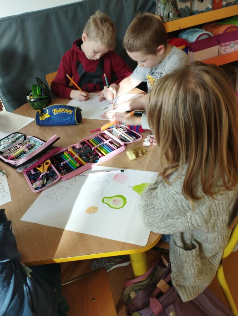 Zdjęcie przedstawia rysujące dzieci. Trójka dzieci siedzi przy drewnianym, sześciokątnym stoliku. Blondynka na pierwszym planie, ubrana w szary sweterek siedzi po prawej stronie stołu. Rysuje na kartce A3 zielone owoce : awokado i gruszkę. Na dalszym planie dwoje chłopców rysuje wspólny rysunek na kartce A3. Jeden z chłopców ubrany jest w szarą koszulkę, a drugi w bordowa bluzę. Chłopiec w szarej koszulce trzyma w ręku zieloną kredkę a chłopiec w bordowej bluzie trzyma w dłoni czerwona kredkę. Na stole leżą piórniki z kolorowymi kredkami. Na dalszym planie widać oparty o szara ścianę zielony materac a po prawej stronie szafkę z kolorowymi zabawkami.