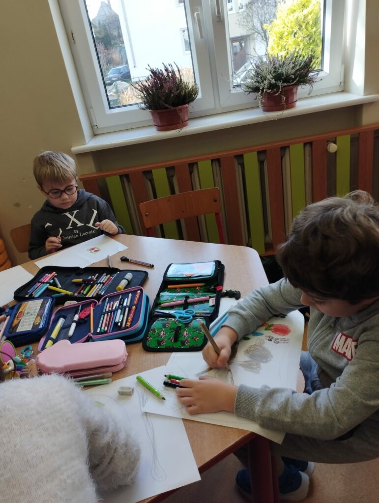 Zdjęcie przedstawia dwóch chłopców rysujących prace plastyczną. Siedzą oni przy brązowym, sześciokątnym stoliku. Chłopiec na pierwszym planie ubrany jest w bezową bluzę, trzyma w ręku brązowa kredkę. Chłopiec siedzący na drugim planie ubrany jest w czarną bluzę z białym wzorem. Nosi okulary. Na stole lezą otwarte piórniki z kolorowym wyposażeniem. Stolik stoi na tle beżowej ściany oraz okna. Za oknem widać szary dom. Na parapecie stoją dwa kolorowe wrzosy w brązowych doniczkach. Kaloryfer pod oknem zakryty jest brązowo- zieloną obudową.