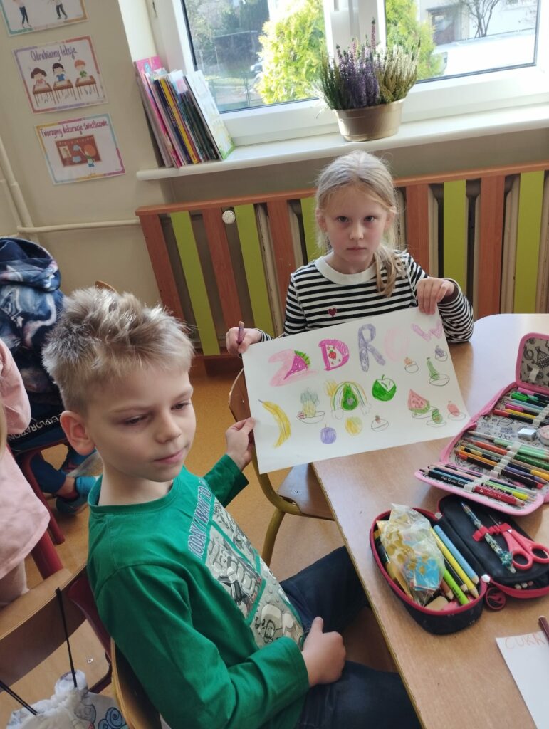 Zdjęcie przedstawia dwoje dzieci siedzących przy brązowym stoliku. Na stoiku lezą kolorowe otwarte piórniki . Chłopiec na pierwszym planie m blond włosy i ubrany jest w zieloną bluzę. Dziewczynka siedząca na dalszym planie ubrana jest w bluzeczkę w biało- czarne paski. Włosy blond ma związane do tyłu. Dziewczynka trzyma w ręku rysunek z kolorowymi owocami i warzywami, który narysowała razem z chłopcem. W tle okno , za którym widać zielony krzew. Na parapecie stoją kolorowe książki i widać kolorowy wrzos w brązowej doniczce.