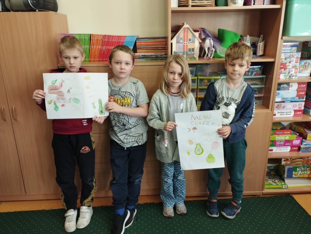 Zdjęcie przedstawia czwórkę dzieci ubranych w kolorowe ubrania i stojących na zielonym dywanie. Za plecami dzieci widać brązowe szafki różnej wysokości , na których leżą kolorowe zabawki książki. Dzieci trzymają w rękach dwie wykonane przez siebie prace plastyczne, które przedstawiają zdrowe warzywa i owoce.