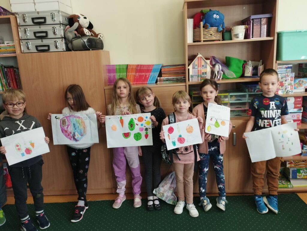 Zdjęcie przedstawia siedmioro dzieci ubranych w kolorowe ubrania i stojących na zielonym dywanie. Za plecami dzieci widać brązowe szafki różnej wysokości , na których leżą kolorowe zabawki książki. Dzieci trzymają w rękach wykonane przez siebie prace plastyczne, które przedstawiają zdrowe warzywa i owoce.