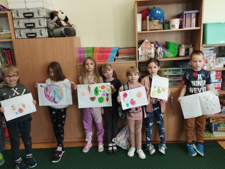 Zdjęcie przedstawia siedmioro dzieci ubranych w kolorowe ubrania i stojących na zielonym dywanie. Za plecami dzieci widać brązowe szafki różnej wysokości , na których leżą kolorowe zabawki książki. Dzieci trzymają w rękach wykonane przez siebie prace plastyczne, które przedstawiają zdrowe warzywa i owoce.